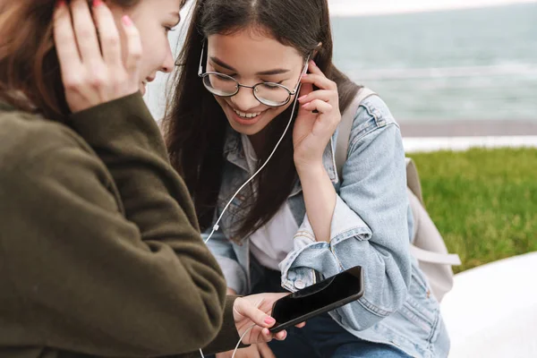 행복한 정서적 젊은 예쁜 친구 여성 학생들은 휴대 전화를 사용하여 이어폰으로 음악을 듣고 야외에서 걷는 학생들. — 스톡 사진