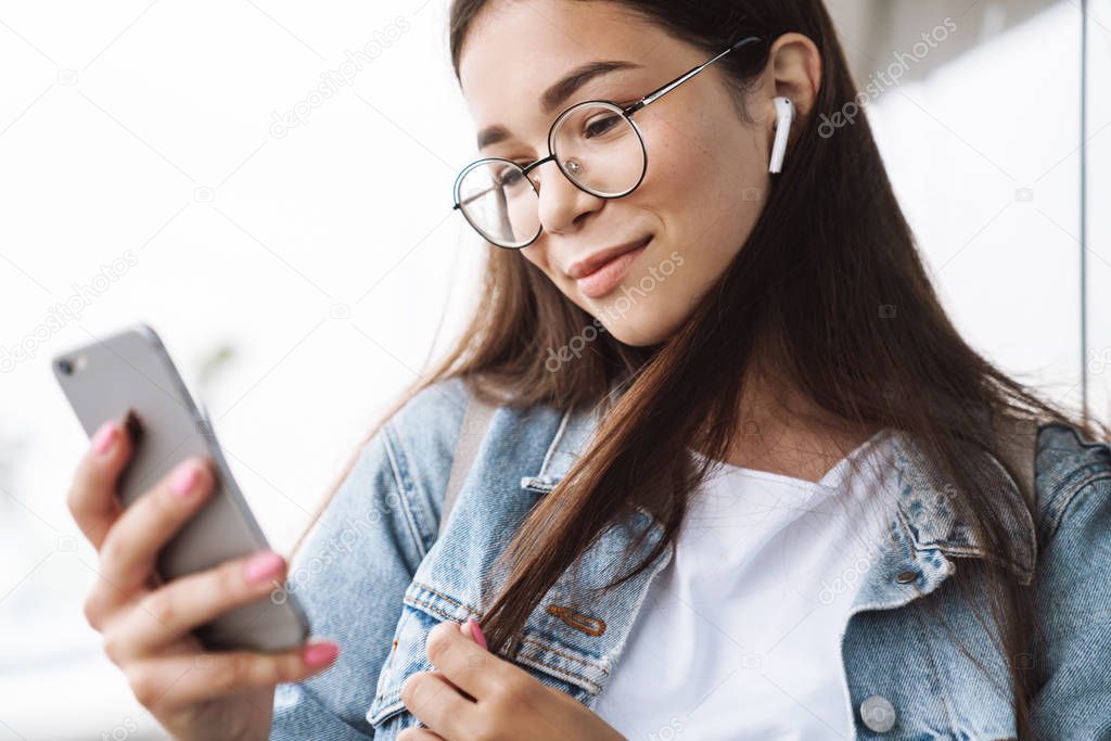 Pleased young pretty woman student wearing eyeglasses walking outdoors resting listening music with earphones using mobile phone chatting.