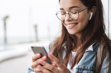 Gülen genç güzel kadın öğrenci cep telefonu sohbet kullanarak kulaklık la müzik dinleyerek açık havada yürüyüş gözlük giyen.