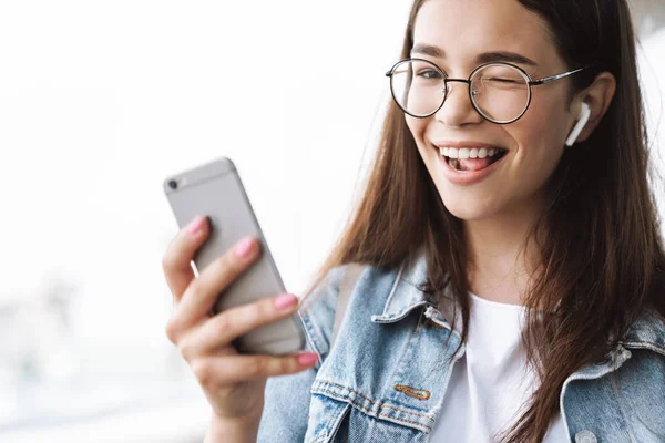 Boldog fiatal szép nő hallgató fárasztó szemüveg gyaloglás kint hallgató zene-val fülhallgató használ mozgatható telefon fog egy selfie. — Stock Fotó