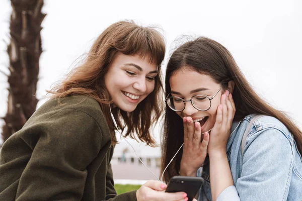 Geschokt jonge mooie vrienden vrouwen studenten wandelen buitenshuis luisteren muziek met oortelefoons met behulp van mobiele telefoon. — Stockfoto