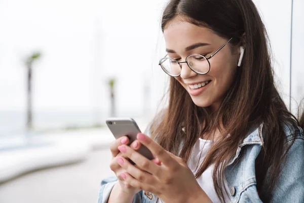 Mosolygó fiatal szép nő hallgató fárasztó szemüvegek gyaloglás kint hallgató zene-val fülhallgató használ mozgatható telefon beszélgető. — Stock Fotó