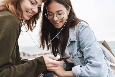 Mutlu duygusal genç güzel arkadaşlar kadın öğrenciler cep telefonu kullanarak kulaklık ile müzik dinlerken açık havada yürüyüş.