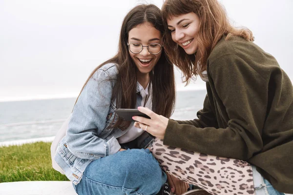 Glad ung vackra vänner kvinnor studenter sitter utomhus med hjälp av mobiltelefon. — Stockfoto
