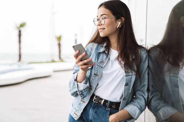 Mosolygó fiatal szép nő hallgató fárasztó szemüvegek gyaloglás kint hallgató zene-val fülhallgató használ mozgatható telefon beszélgető. Látszó félre. — Stock Fotó