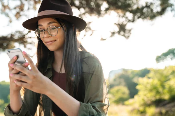 Fotó a vonzó barna nő visel stílusos kalap és szemüvegek segítségével cellphone járás közben a zöld parkban — Stock Fotó