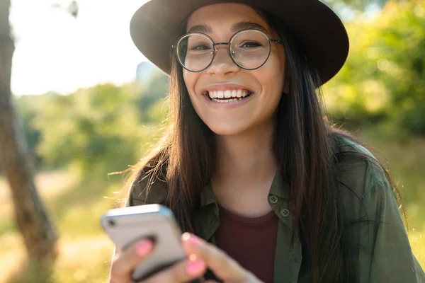 Fénykép-ból vonzó nevető asszony fárasztó elegáns kalap és szemüvegek használ cellphone rövid idő gyaloglás-ban zöld Park — Stock Fotó