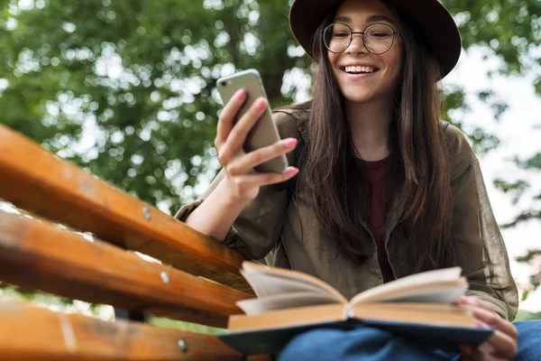 Foto einer glücklichen Frau mit langen dunklen Haaren, die Hut und Brille mit Smartphone trägt, während sie auf einer Bank im grünen Park ein Buch liest — Stockfoto