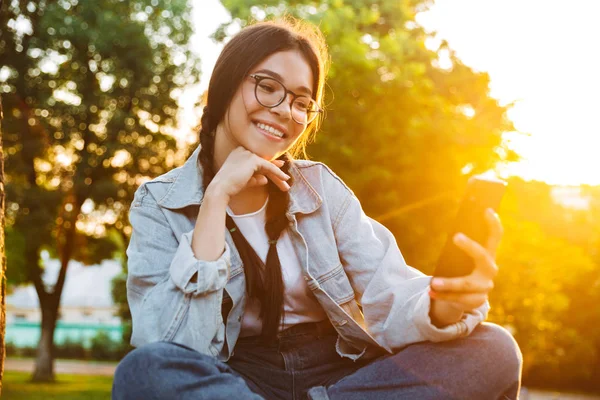 Sorridente allegro carino giovane studentessa indossa occhiali da vista seduto sulla panchina all'aperto nel parco naturale con bella luce del sole utilizzando il telefono cellulare . — Foto Stock