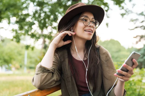 Fotó szép nő viselt kalap és szemüvegek segítségével a fülhallgató és okostelefon zöld Park — Stock Fotó