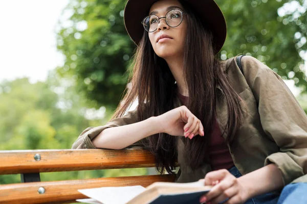 坐在绿色公园的长椅上，集中的妇女戴着帽子和眼镜看书的照片 — 图库照片
