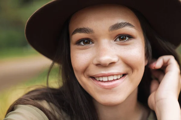 Foto primo piano di allegra bella donna che indossa cappello sorridente alla macchina fotografica mentre seduto su erba all'aperto — Foto Stock