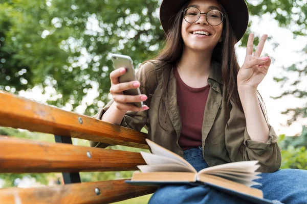Fotó a fiatal nő viselt kalap és szemüvegek segítségével okostelefon és mutatja béke jel padon zöld parkban — Stock Fotó