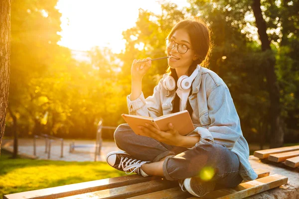 Όνειρα σκέψης νεαρό μαθητή κορίτσι φορώντας γυαλιά κάθονται σε εξωτερικούς χώρους στο φυσικό πάρκο ακούγοντας μουσική με τα ακουστικά γράφοντας σημειώσεις. — Φωτογραφία Αρχείου