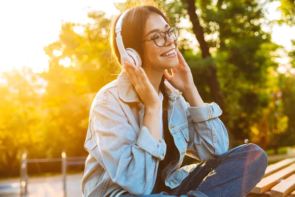Agradable joven adolescente feliz estudiante sentado al aire libre en el hermoso parque verde escuchando música con auriculares . —  Fotos de Stock