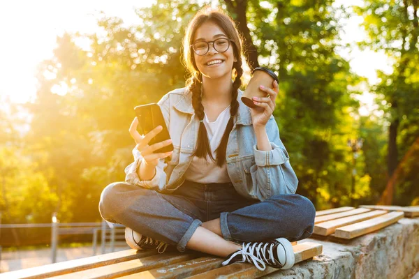 Boldog aranyos fiatal tanuló lány visel szemüvegek ül a szabadban a természetvédelmi parkban mobiltelefon fogyasztása kávét. — Stock Fotó
