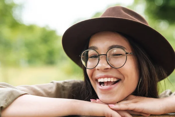 Foto closeup de jovem mulher vestindo chapéu e óculos sorrindo com a boca aberta e piscando enquanto sentado no banco — Fotografia de Stock