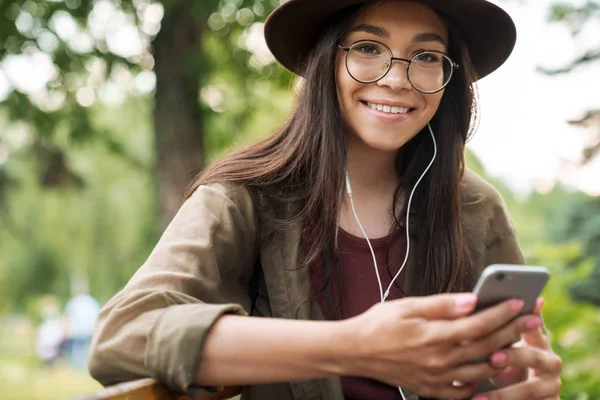 Foto de una mujer sonriente que usa sombrero y anteojos usando auriculares y teléfonos inteligentes en el parque verde — Foto de Stock