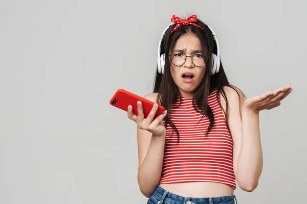 Спантеличена молода незадоволена дівчина-підліток, одягнена в яскраво-червону футболку, використовуючи мобільний телефон ізольована на сірому фоні стіни . — стокове фото