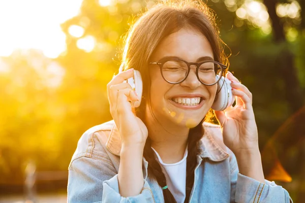 Agradable joven adolescente feliz estudiante sentado al aire libre en el hermoso parque verde escuchando música con auriculares . —  Fotos de Stock