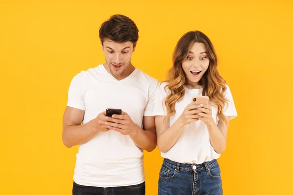 Retrato de una hermosa pareja hombre y mujer en camisetas básicas de pie juntos mientras sostiene los teléfonos inteligentes — Foto de Stock