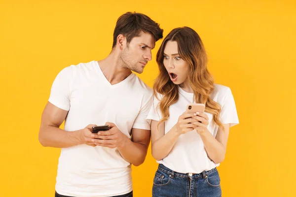 Foto de casal perplexo homem e mulher em camisetas básicas segurando e espreitando telefones celulares — Fotografia de Stock