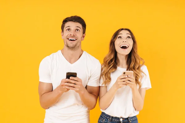 Porträtt av förvånade par man och kvinna i grundläggande t-shirts ser uppåt tillsammans på copyspace medan du håller smartphones — Stockfoto