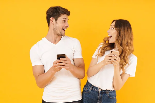 Retrato de una hermosa pareja de hombres y mujeres en camisetas básicas mirándose mientras sostienen teléfonos inteligentes — Foto de Stock