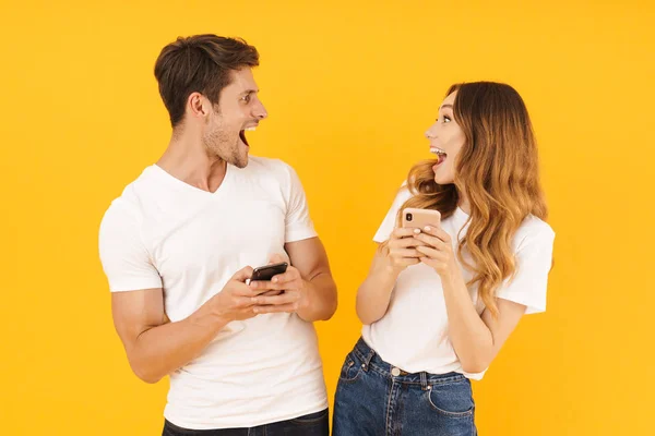Portretul unui cuplu atractiv bărbat și femeie în tricouri de bază care se uită unul la celălalt în timp ce dețin smartphone-uri — Fotografie, imagine de stoc