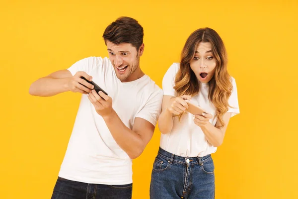 Retrato de casal feliz homem e mulher em camisetas básicas de pé juntos enquanto joga jogos de vídeo em smartphones — Fotografia de Stock