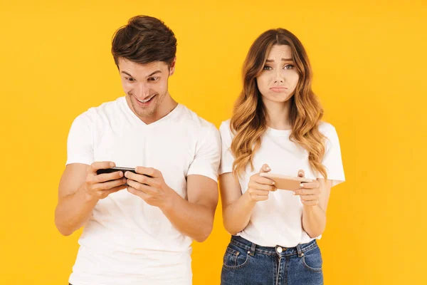 Retrato de pareja joven hombre y mujer en camisetas básicas de pie juntos mientras juegan videojuegos en teléfonos inteligentes — Foto de Stock