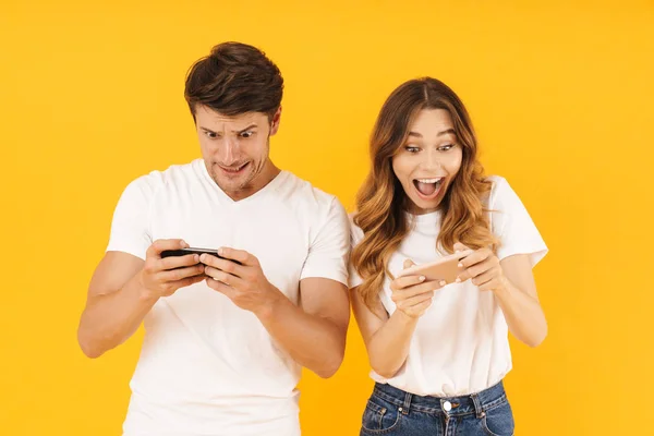 Porträtt av Joyful par man och kvinna i grundläggande t-shirts stående tillsammans medan du spelar TV-spel på smartphones — Stockfoto