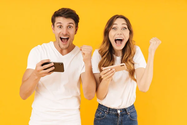 Porträtt av förvånad par man och kvinna i grundläggande t-shirts stående tillsammans medan du spelar video spel på smartphones — Stockfoto