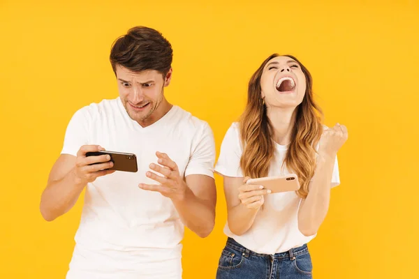 Porträtt av förtjust par man och kvinna i grundläggande t-shirts stående tillsammans medan du spelar video spel på smartphones — Stockfoto