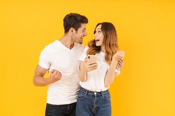Портрет довольной пары мужчины и женщины в основных футболках, смотрящих друг на друга, держа в руках смартфон — стоковое фото