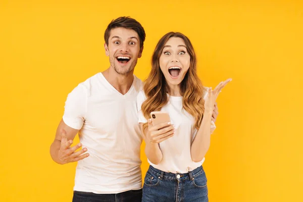Retrato de pareja excitada hombre y mujer en camisetas básicas regocijándose mientras están de pie junto con el teléfono inteligente — Foto de Stock