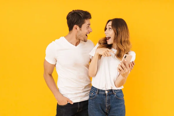 Портрет красивой пары мужчины и женщины в основных футболках, смотрящих друг на друга, держа в руках смартфон — стоковое фото