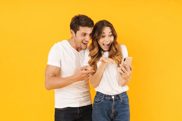 Retrato de pareja asombrada hombre y mujer en camisetas básicas regocijándose mientras usan juntos el teléfono inteligente — Foto de Stock