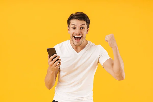 Feliz emocionado joven sorprendido en casual camiseta blanca utilizando el teléfono móvil aislado sobre fondo amarillo . — Foto de Stock