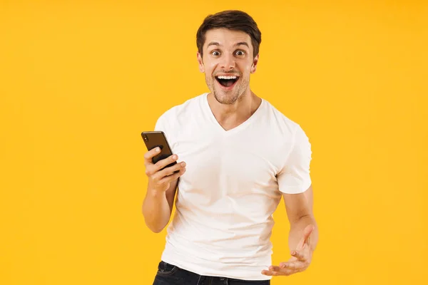 Feliz emocionado joven sorprendido en casual camiseta blanca utilizando el teléfono móvil aislado sobre fondo amarillo . — Foto de Stock