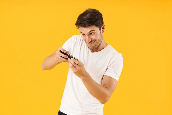 Jovem nervoso em casual branco t-shirt jogar jogos por telefone celular isolado sobre fundo amarelo . — Fotografia de Stock