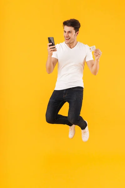 欢快的兴奋惊讶年轻男子在休闲白色T恤用手机跳隔离在黄色背景持有信用卡. — 图库照片