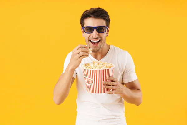 Молодий чоловік у повсякденній білій футболці їсть попкорн в 3d окулярах дивитися фільм ізольовано на жовтому тлі . — стокове фото