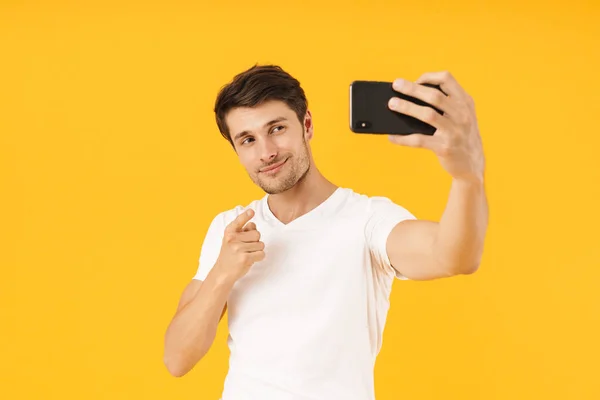 Arrogant young pleased man in casual white t-shirt take a selfie by mobile phone isolated over yellow background. — Stock Photo, Image