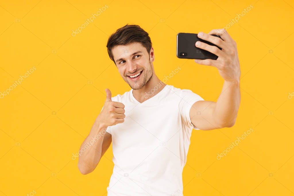 Happy young cheerful man in casual white t-shirt take a selfie by mobile phone isolated over yellow background.