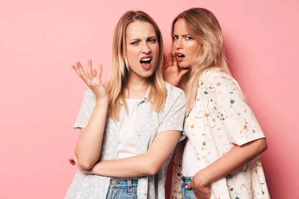 Ontevreden verward mooie blondjes vrouwen vrienden poseren geïsoleerd over roze muur achtergrond praten met elkaar. — Stockfoto