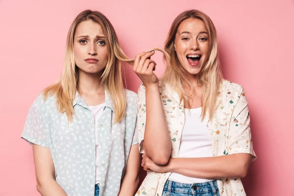 Happy woman holding hair of her sad pretty blonde women friend sister posing isolated over pink wall background. — Stock Photo, Image