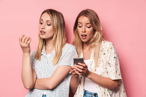 Muchacha conmocionada usando el teléfono móvil que muestra la pantalla a su indiferente teléfono amigo mujer aislado sobre fondo de pared rosa . — Foto de Stock