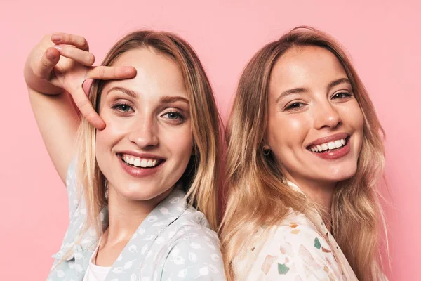 Веселые, довольные, красивые блондинки подруги позируют изолированно на фоне розовой стены, показывая жест мира . — стоковое фото