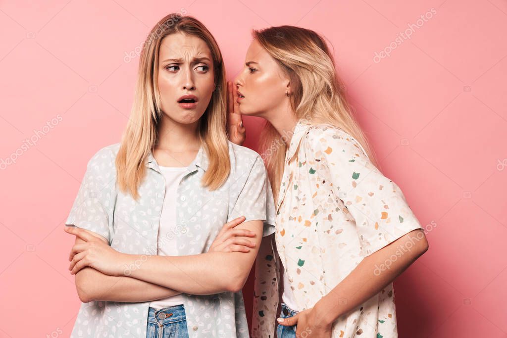 Negative beautiful blondes women friends posing isolated over pink wall background talking with each other gossiping.